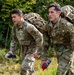 Two U.S. Army soldiers in Operational Camouflage Pattern (OCP) uniforms are conducting a ruck march through a wooded area. Both soldiers are carrying large military rucksacks and holding water bottles, suggesting an endurance training exercise. The soldier on the right displays a Ranger tab and what appears to be a 10th Mountain Division patch.