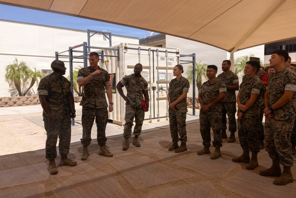 A group of U.S. Marines in woodland MARPAT camouflage uniforms are gathered in an outdoor covered area, appearing to receive a briefing or instruction from a senior Marine. The setting appears to be a military installation in a warm, arid climate, with a shipping container and pull-up bar rack visible in the background. The group includes both male and female Marines standing in an attentive formation.