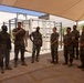 A group of U.S. Marines in woodland MARPAT camouflage uniforms are gathered in an outdoor covered area, appearing to receive a briefing or instruction from a senior Marine. The setting appears to be a military installation in a warm, arid climate, with a shipping container and pull-up bar rack visible in the background. The group includes both male and female Marines standing in an attentive formation.