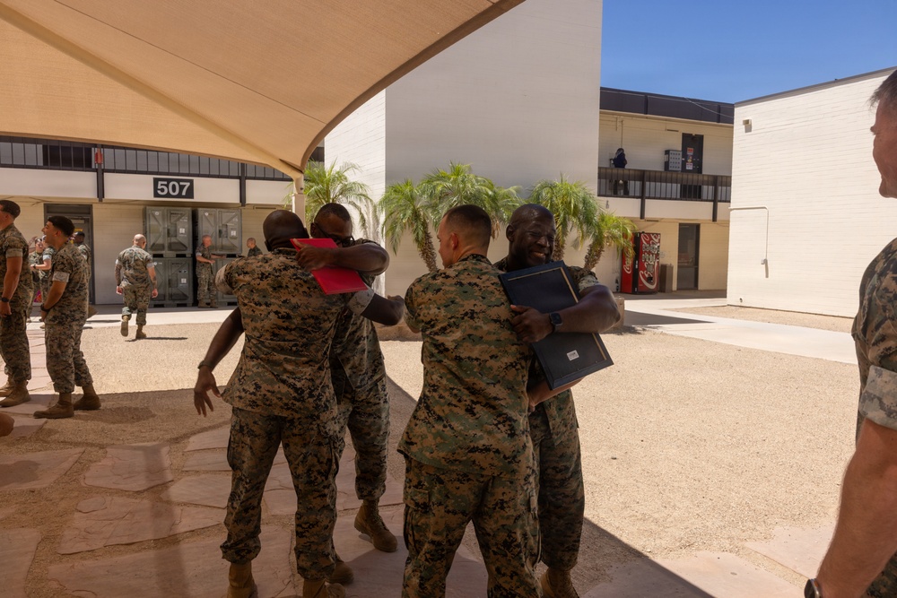Military personnel in MARPAT camouflage uniforms are gathered in an outdoor courtyard area, with several individuals embracing in what appears to be a celebratory or farewell occasion. Two pairs of service members are hugging in the foreground, with one individual holding a dark blue folder or certificate binder. The setting appears to be a U.S. Marine Corps installation, identifiable by building number 507 visible in the background.