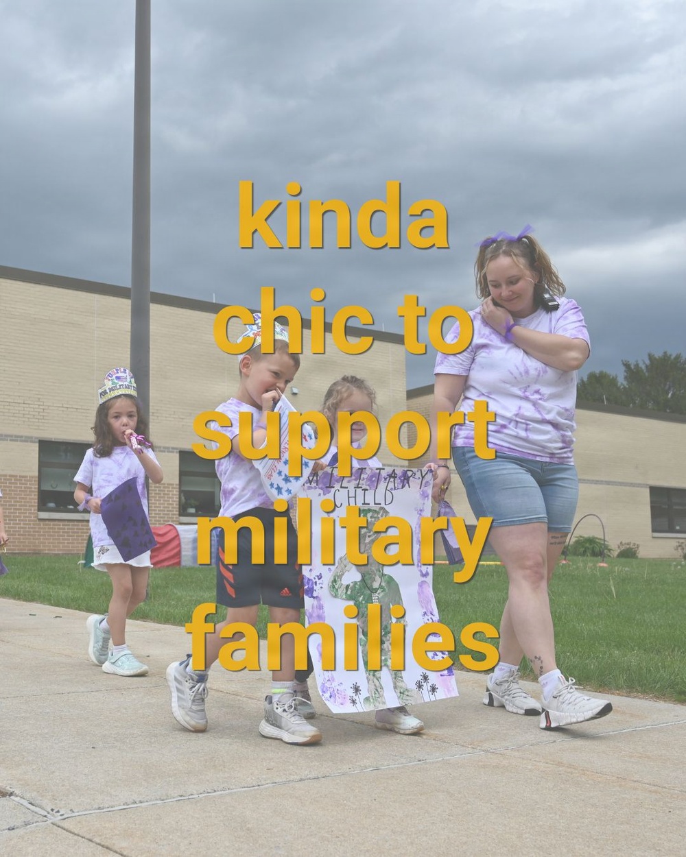 A group of children and an adult woman are participating in a military family support event, walking along a sidewalk outside a brick building. The participants are wearing matching purple tie-dye shirts, and two children are holding a hand-decorated sign reading 'Military Child.' Overlaid text reads 'kinda chic to support military families.'