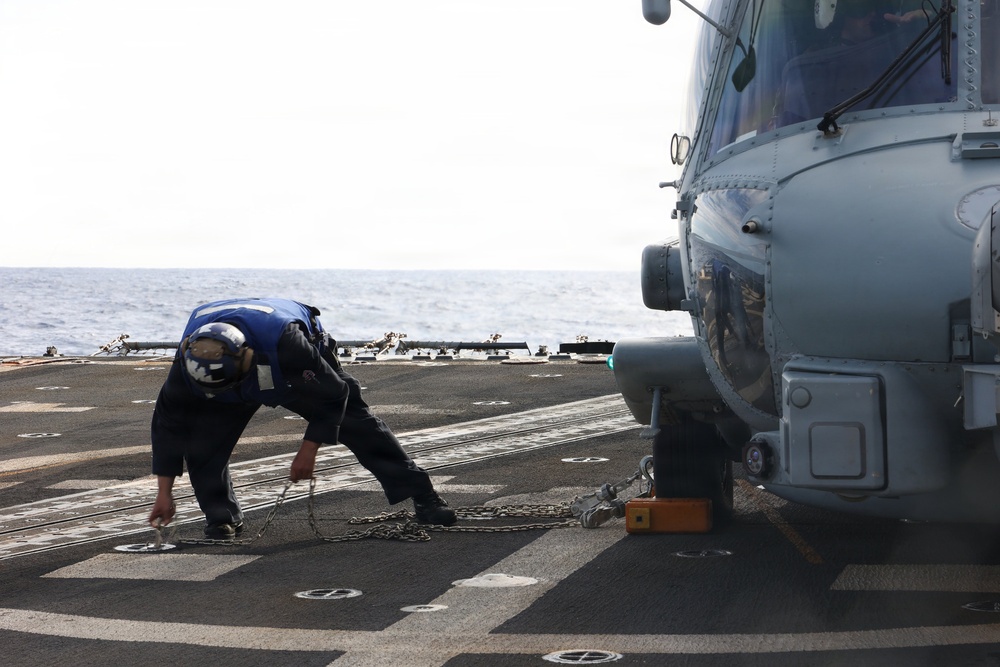 A naval crew member in blue coveralls and a helmet is working on a ship's flight deck, handling chains used to secure a military helicopter. The helicopter, painted in gray, is chocked and chained to the deck of what appears to be a naval vessel at sea. The ocean horizon is visible in the background, confirming an open-water operational environment.