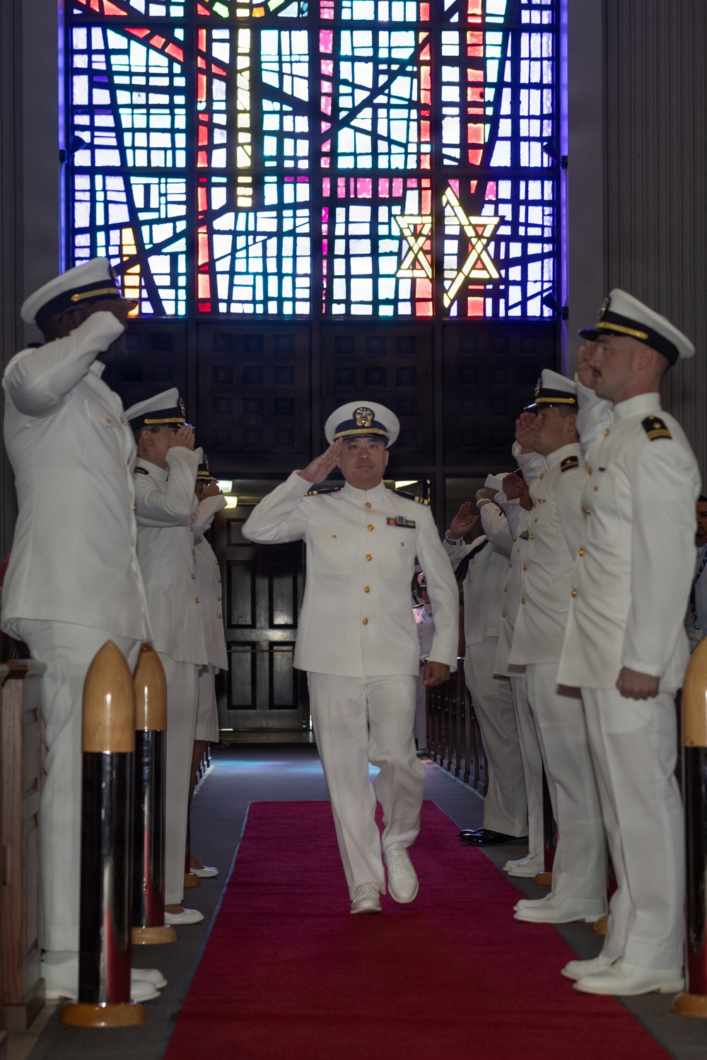 A military officer in white dress uniform walks down a red carpet aisle while fellow officers on both sides render salutes in a formal ceremony. The setting appears to be a chapel or ceremonial hall, evidenced by a large colorful stained glass window in the background. This appears to be a traditional military 'arch of sabers' or honor guard passage ceremony.