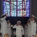 A military officer in white dress uniform walks down a red carpet aisle while fellow officers on both sides render salutes in a formal ceremony. The setting appears to be a chapel or ceremonial hall, evidenced by a large colorful stained glass window in the background. This appears to be a traditional military 'arch of sabers' or honor guard passage ceremony.