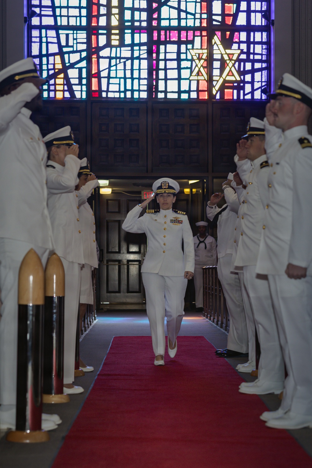A U.S. Navy officer in white dress uniform walks down a red carpet through a sideboy honors corridor, saluting while fellow officers on both sides render salutes. The ceremony appears to be a formal military event such as a change of command or retirement ceremony taking place inside a chapel or ceremonial hall. A large colorful stained glass window featuring Star of David motifs is prominently visible in the background.