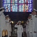 A U.S. Navy officer in white dress uniform walks down a red carpet through a sideboy honors corridor, saluting while fellow officers on both sides render salutes. The ceremony appears to be a formal military event such as a change of command or retirement ceremony taking place inside a chapel or ceremonial hall. A large colorful stained glass window featuring Star of David motifs is prominently visible in the background.