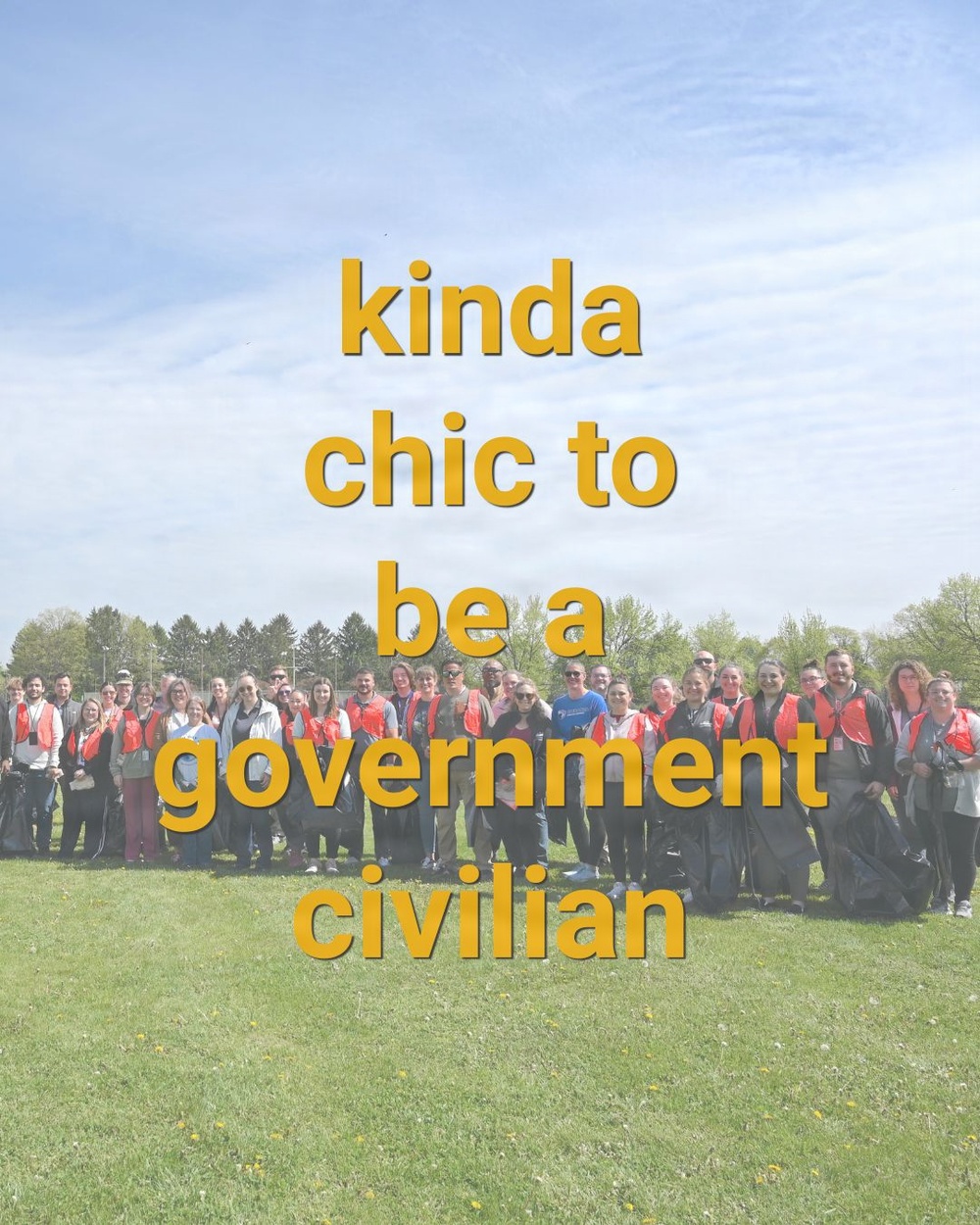 A large group of approximately 30-40 people pose together outdoors on a grassy field under a partly cloudy sky. Many participants are wearing matching red and black vests or jackets, suggesting an organized group activity or program. The image is overlaid with bold yellow text reading 'kinda chic to be a government civilian.'