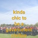 A large group of approximately 30-40 people pose together outdoors on a grassy field under a partly cloudy sky. Many participants are wearing matching red and black vests or jackets, suggesting an organized group activity or program. The image is overlaid with bold yellow text reading 'kinda chic to be a government civilian.'
