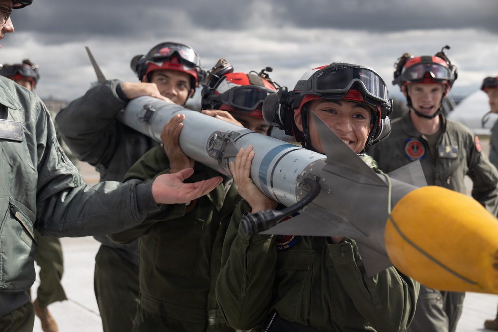 Military personnel in flight suits and red-helmeted gear are collectively carrying a missile or air-to-air munition on an airfield tarmac. The team appears to be conducting weapons loading training or a competition drill, with multiple crew members working together to transport the ordnance. A yellow safety cap is visible on the missile's rear fin assembly.