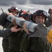 Military personnel in flight suits and red-helmeted gear are collectively carrying a missile or air-to-air munition on an airfield tarmac. The team appears to be conducting weapons loading training or a competition drill, with multiple crew members working together to transport the ordnance. A yellow safety cap is visible on the missile's rear fin assembly.