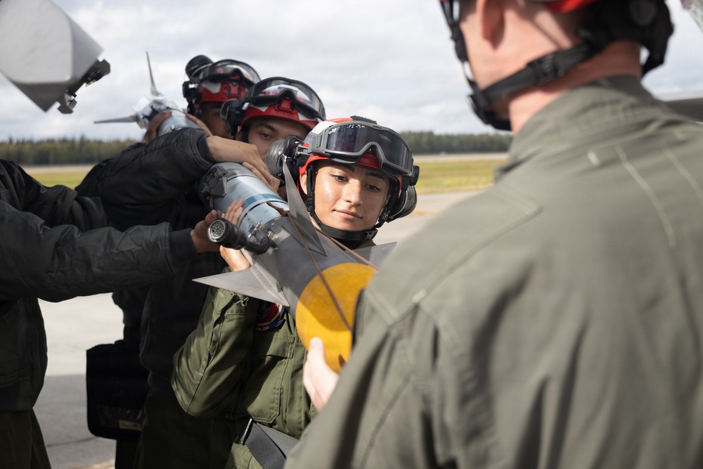 Military personnel in flight suits and red helmets with protective goggles are gathered on an airfield tarmac, handling what appears to be a missile or ordnance component. A person in the foreground with their back to the camera appears to be instructing or supervising the group. The setting appears to be a military air base with aircraft and tree line visible in the background.