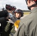 Military personnel in flight suits and red helmets with protective goggles are gathered on an airfield tarmac, handling what appears to be a missile or ordnance component. A person in the foreground with their back to the camera appears to be instructing or supervising the group. The setting appears to be a military air base with aircraft and tree line visible in the background.