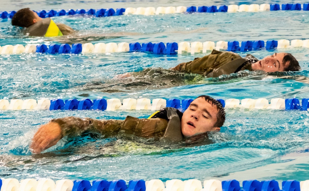 Three individuals are swimming in an indoor lap pool with blue and white lane dividers. At least two of the swimmers in the foreground are wearing what appears to be military-style camouflage uniforms and life vests/flotation devices while swimming, suggesting a military water survival training exercise. The swimmers are using a backstroke or side-stroke technique while fully clothed.