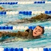 Three individuals are swimming in an indoor lap pool with blue and white lane dividers. At least two of the swimmers in the foreground are wearing what appears to be military-style camouflage uniforms and life vests/flotation devices while swimming, suggesting a military water survival training exercise. The swimmers are using a backstroke or side-stroke technique while fully clothed.