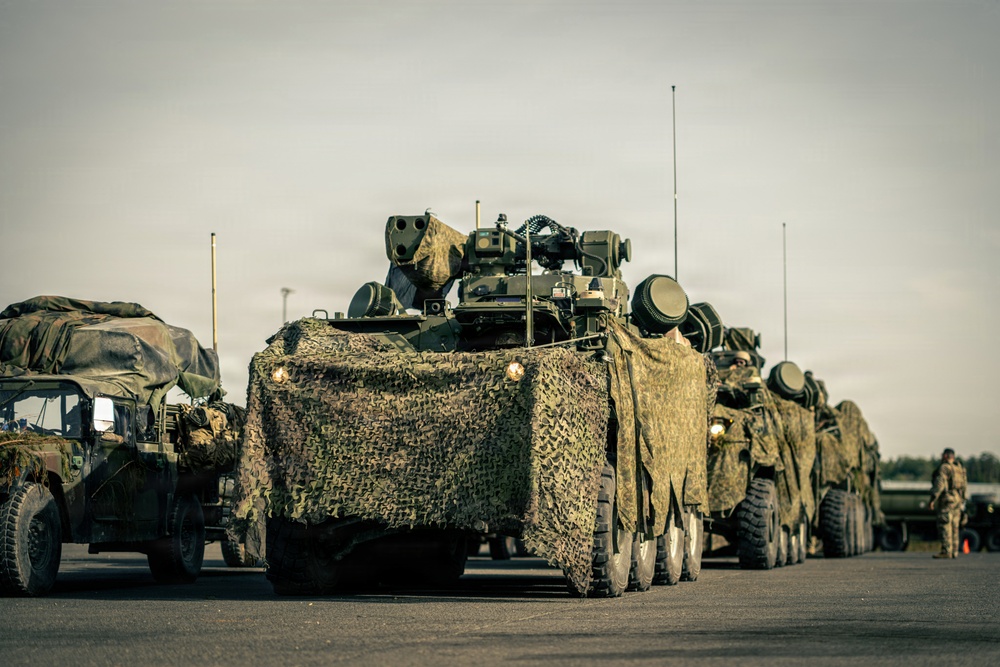 A military convoy of wheeled armored vehicles is staged or moving along a paved road or tarmac. The lead vehicle is draped in camouflage netting and appears to be equipped with a weapons or sensor system on top. At least one soldier is visible in the background on the right side of the frame.