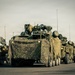 A military convoy of wheeled armored vehicles is staged or moving along a paved road or tarmac. The lead vehicle is draped in camouflage netting and appears to be equipped with a weapons or sensor system on top. At least one soldier is visible in the background on the right side of the frame.