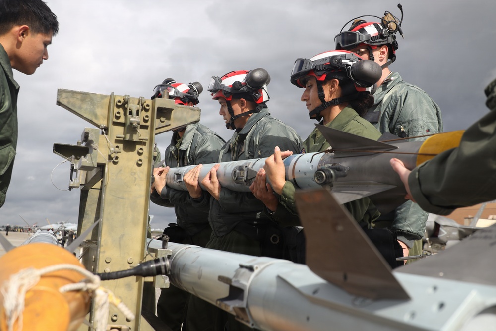 Military personnel in flight suits and red-and-white helmets with goggles are handling what appears to be an air-to-air missile on a weapons loading cart on a flight line. The group appears to be conducting weapons loading or maintenance training on aircraft ordnance. The setting is an outdoor airfield with overcast skies and additional aircraft visible in the background.