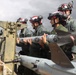 Military personnel in flight suits and red-and-white helmets with goggles are handling what appears to be an air-to-air missile on a weapons loading cart on a flight line. The group appears to be conducting weapons loading or maintenance training on aircraft ordnance. The setting is an outdoor airfield with overcast skies and additional aircraft visible in the background.