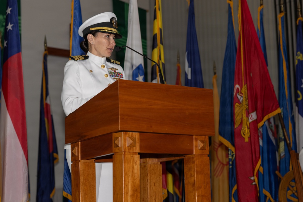 A female U.S. Navy officer in white dress uniform is speaking at a wooden podium with a microphone. She is surrounded by numerous military and state flags in what appears to be an official ceremony or formal military event. Her uniform displays gold shoulder boards indicating senior officer rank, along with multiple ribbons and insignia.