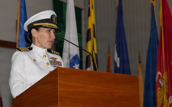 A female U.S. Navy officer in white dress uniform is speaking at a wooden podium with a microphone. She is surrounded by numerous military and state flags in what appears to be an official ceremony or formal military event. Her uniform displays gold shoulder boards indicating senior officer rank, along with multiple ribbons and insignia.
