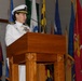 A female U.S. Navy officer in white dress uniform is speaking at a wooden podium with a microphone. She is surrounded by numerous military and state flags in what appears to be an official ceremony or formal military event. Her uniform displays gold shoulder boards indicating senior officer rank, along with multiple ribbons and insignia.