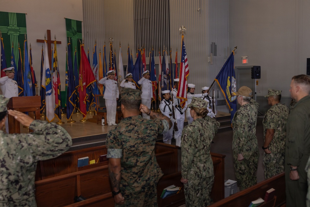 A military ceremony is taking place inside what appears to be a chapel or auditorium on a military installation. Navy personnel in white dress uniforms are on stage with a color guard presenting flags, while a mixed audience of Navy and Marine Corps personnel in camouflage uniforms stand at attention and salute. Multiple state and military flags are displayed on the stage alongside a Christian cross.