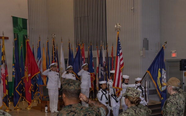A military ceremony is taking place inside what appears to be a chapel or auditorium on a military installation. Navy personnel in white dress uniforms are on stage with a color guard presenting flags, while a mixed audience of Navy and Marine Corps personnel in camouflage uniforms stand at attention and salute. Multiple state and military flags are displayed on the stage alongside a Christian cross.
