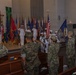 A military ceremony is taking place inside what appears to be a chapel or auditorium on a military installation. Navy personnel in white dress uniforms are on stage with a color guard presenting flags, while a mixed audience of Navy and Marine Corps personnel in camouflage uniforms stand at attention and salute. Multiple state and military flags are displayed on the stage alongside a Christian cross.