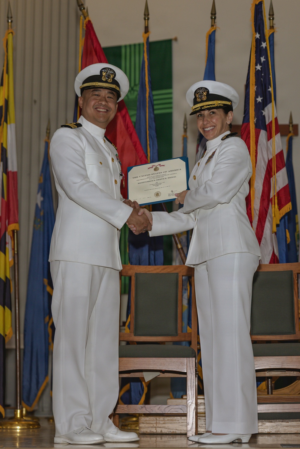 Two U.S. Navy officers in white dress uniforms are shaking hands while jointly holding a certificate during a formal military ceremony. The certificate is visibly titled 'The United States of America' and references a 'Meritorious Service Medal.' The setting includes multiple flags, including the U.S. flag, arranged behind them on a stage.