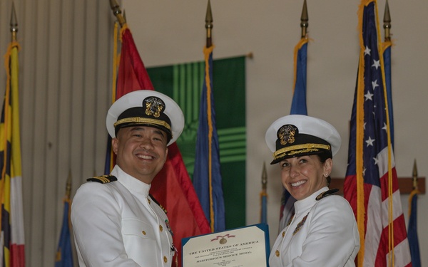 Two U.S. Navy officers in white dress uniforms are shaking hands while jointly holding a certificate during a formal military ceremony. The certificate is visibly titled 'The United States of America' and references a 'Meritorious Service Medal.' The setting includes multiple flags, including the U.S. flag, arranged behind them on a stage.