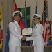 Two U.S. Navy officers in white dress uniforms are shaking hands while jointly holding a certificate during a formal military ceremony. The certificate is visibly titled 'The United States of America' and references a 'Meritorious Service Medal.' The setting includes multiple flags, including the U.S. flag, arranged behind them on a stage.