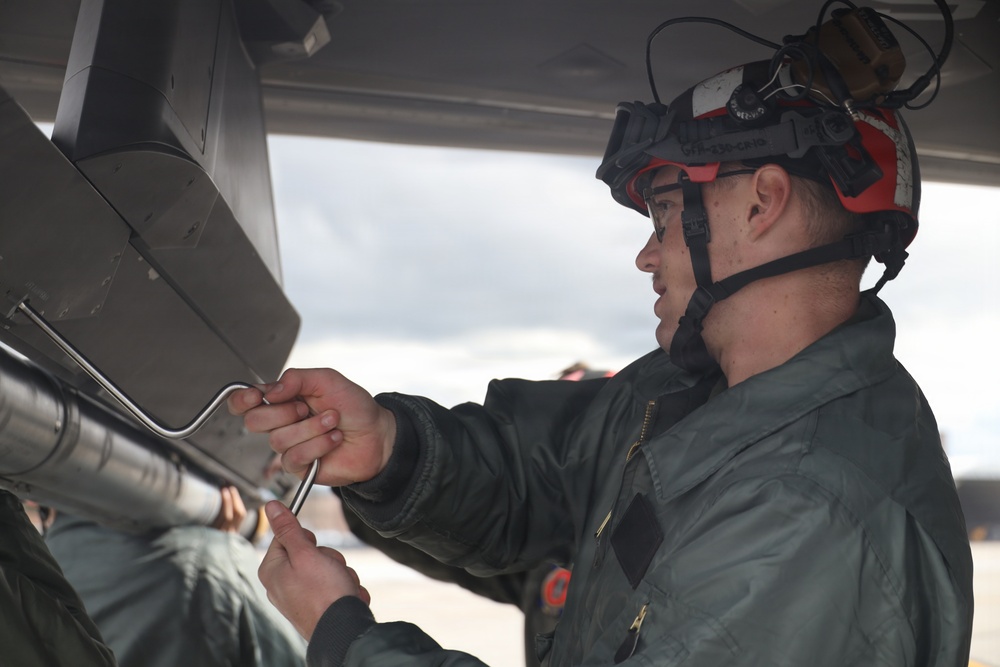 A military aviation technician or crew member wearing a red and white flight helmet with attached night vision goggle mount is performing maintenance or inspection work on an aircraft component. The individual is dressed in a sage/olive green flight suit and is manipulating what appears to be a safety pin or locking wire on an aircraft system. The setting appears to be an outdoor flight line or tarmac under overcast skies.