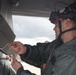A military aviation technician or crew member wearing a red and white flight helmet with attached night vision goggle mount is performing maintenance or inspection work on an aircraft component. The individual is dressed in a sage/olive green flight suit and is manipulating what appears to be a safety pin or locking wire on an aircraft system. The setting appears to be an outdoor flight line or tarmac under overcast skies.