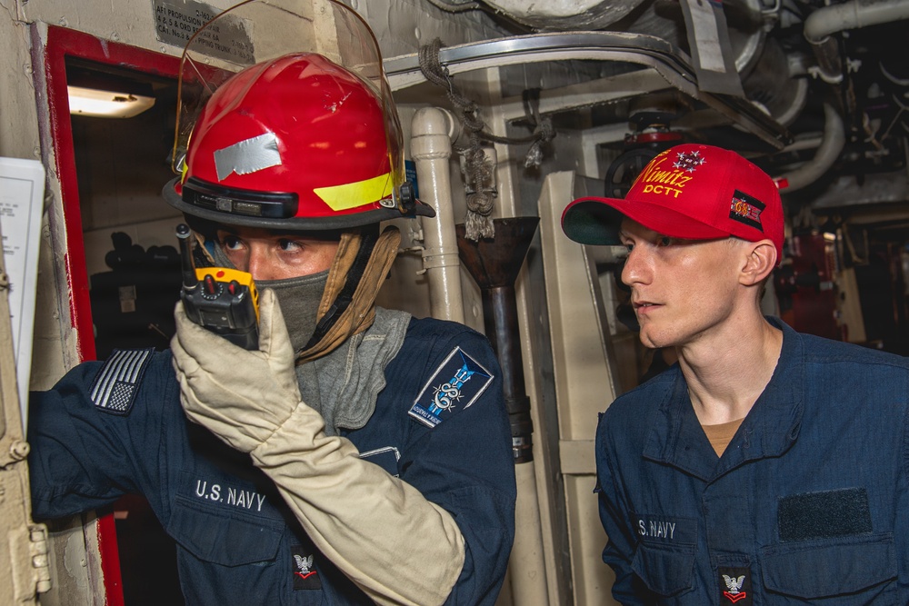 Two U.S. Navy sailors are photographed inside what appears to be a shipboard compartment, likely during a damage control training exercise. The sailor on the left is wearing firefighting gear including a red helmet and protective gloves, and is holding a radio to communicate. The sailor on the right is wearing a red 'Nimitz DCTT' cap, indicating involvement with the USS Nimitz Damage Control Training Team.