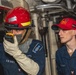 Two U.S. Navy sailors are photographed inside what appears to be a shipboard compartment, likely during a damage control training exercise. The sailor on the left is wearing firefighting gear including a red helmet and protective gloves, and is holding a radio to communicate. The sailor on the right is wearing a red 'Nimitz DCTT' cap, indicating involvement with the USS Nimitz Damage Control Training Team.