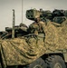 The image shows military personnel operating what appears to be a Stryker armored fighting vehicle draped with camouflage netting. One soldier is visible in the turret hatch area, appearing to look at a device, while another soldier is partially visible in the foreground. The scene suggests a military exercise or operational staging area.