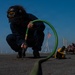 A U.S. Navy sailor assigned to USS Nimitz (CVN-68) is crouched on the flight deck performing maintenance work using a pneumatic or powered tool connected to a green hose. The image is taken from a low ground-level perspective, with the green hose extending toward the camera in the foreground. An American flag is visible on a mast in the background under a clear blue sky.