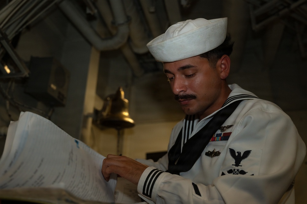 A U.S. Navy sailor in dress white uniform is reviewing documents aboard what appears to be a naval vessel. The sailor wears a traditional white Dixie cup cover and displays several ribbons and insignia on his uniform. The setting appears to be an interior compartment of a ship, with visible pipes and a brass bell in the background.