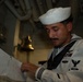 A U.S. Navy sailor in dress white uniform is reviewing documents aboard what appears to be a naval vessel. The sailor wears a traditional white Dixie cup cover and displays several ribbons and insignia on his uniform. The setting appears to be an interior compartment of a ship, with visible pipes and a brass bell in the background.