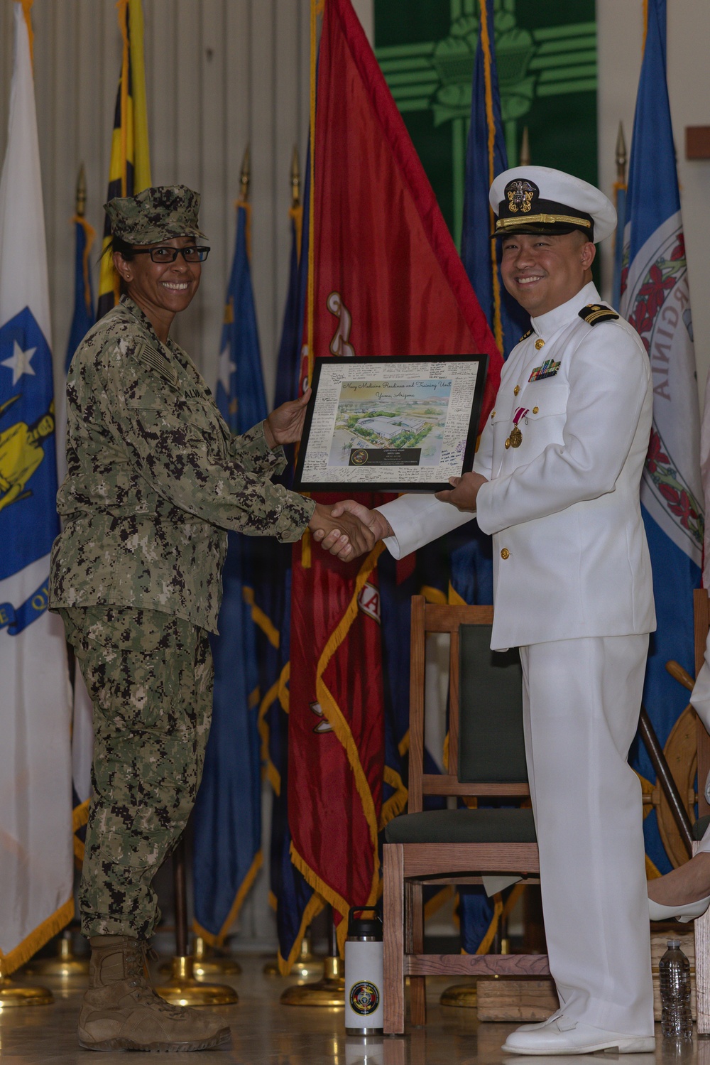 Two U.S. Navy personnel are shaking hands during a formal military ceremony, with one presenting a framed plaque or award to the other. The individual on the left is wearing Navy Working Uniform (NWU) camouflage with a name tag reading 'ALM...' and a single star insignia, while the individual on the right is wearing the formal Navy Service Dress White uniform with officer's cover, medals, and gold buttons. Multiple military and state flags are displayed in the background.