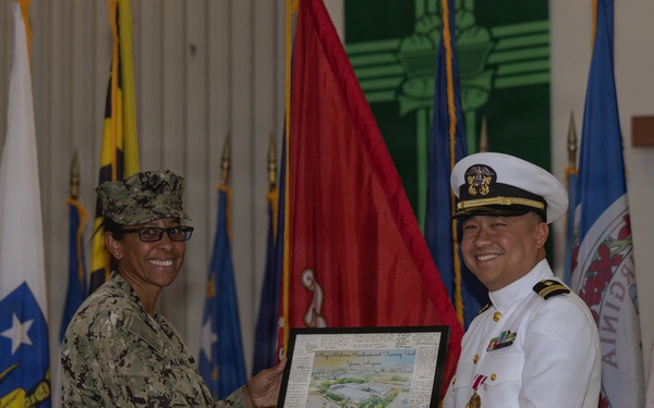 Two U.S. Navy personnel are shaking hands during a formal military ceremony, with one presenting a framed plaque or award to the other. The individual on the left is wearing Navy Working Uniform (NWU) camouflage with a name tag reading 'ALM...' and a single star insignia, while the individual on the right is wearing the formal Navy Service Dress White uniform with officer's cover, medals, and gold buttons. Multiple military and state flags are displayed in the background.
