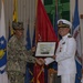 Two U.S. Navy personnel are shaking hands during a formal military ceremony, with one presenting a framed plaque or award to the other. The individual on the left is wearing Navy Working Uniform (NWU) camouflage with a name tag reading 'ALM...' and a single star insignia, while the individual on the right is wearing the formal Navy Service Dress White uniform with officer's cover, medals, and gold buttons. Multiple military and state flags are displayed in the background.