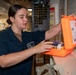 A young female military service member in navy blue utility uniform is accessing an orange emergency or first aid kit mounted near a whiteboard. The setting appears to be the interior of a naval vessel, indicated by the industrial surroundings, metal fixtures, and military-style equipment. She appears to be inspecting or retrieving items from the orange case.