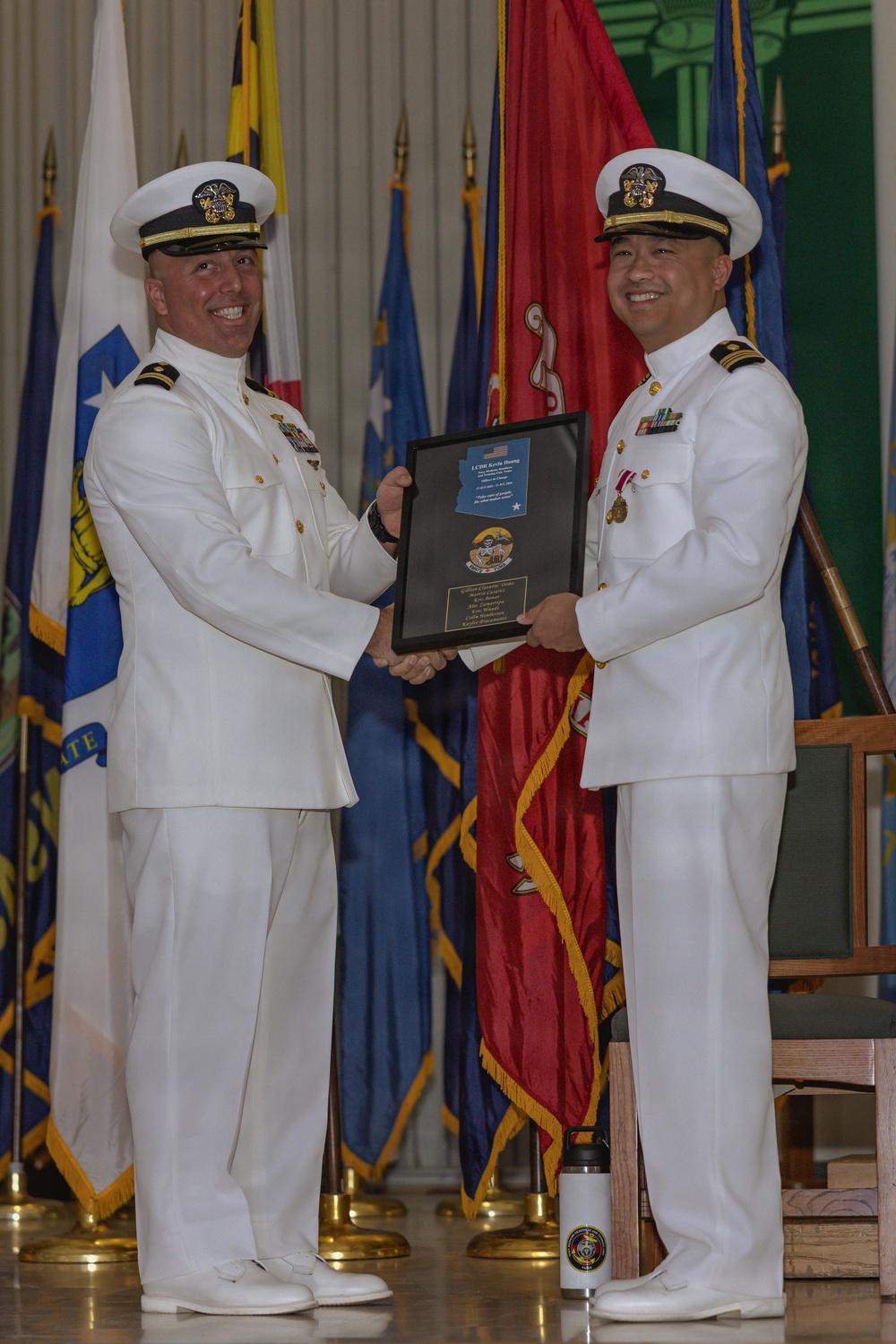 Two U.S. Navy officers in white dress uniforms are participating in a formal military ceremony, jointly holding a framed plaque or award. The event appears to be a change of command, retirement, or recognition ceremony, taking place in a formal hall decorated with multiple military and state flags. The officer on the right is wearing service ribbons and a medal, suggesting he is the recipient of the award.