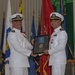 Two U.S. Navy officers in white dress uniforms are participating in a formal military ceremony, jointly holding a framed plaque or award. The event appears to be a change of command, retirement, or recognition ceremony, taking place in a formal hall decorated with multiple military and state flags. The officer on the right is wearing service ribbons and a medal, suggesting he is the recipient of the award.