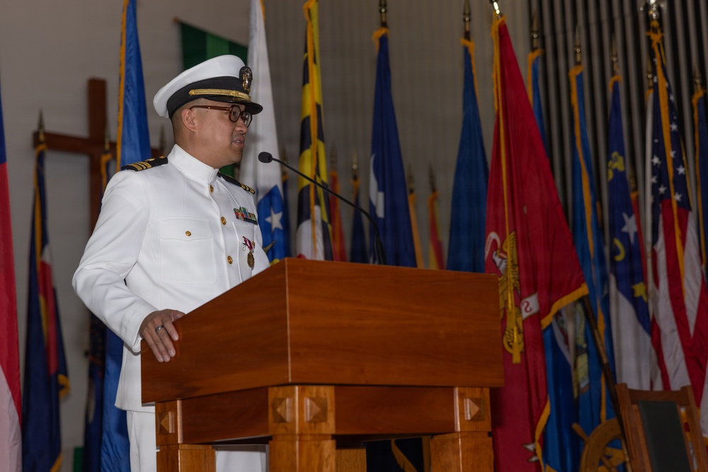 A naval officer in white dress uniform is speaking at a wooden podium during what appears to be a formal military ceremony. Multiple flags are displayed in the background, including what appear to be U.S. military branch flags and state or territorial flags. The setting suggests an official change of command, retirement, or commissioning ceremony.