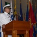 A naval officer in white dress uniform is speaking at a wooden podium during what appears to be a formal military ceremony. Multiple flags are displayed in the background, including what appear to be U.S. military branch flags and state or territorial flags. The setting suggests an official change of command, retirement, or commissioning ceremony.