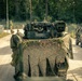 A military convoy is moving along a gravel road through a forested area. The lead vehicle is heavily draped with camouflage netting and natural vegetation for concealment, with military equipment visible on top. Additional military vehicles and personnel on foot are visible in the background.