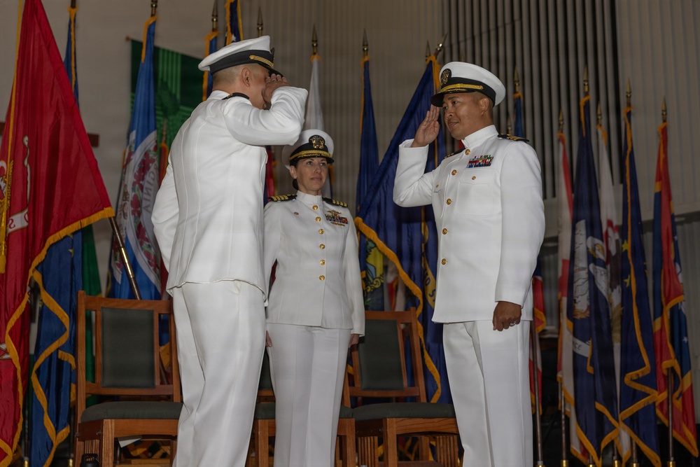 Three U.S. Navy officers in white dress uniforms are participating in a formal military ceremony, likely a change of command. Two officers are exchanging salutes while a third officer, a woman with captain or admiral insignia, stands between them as a presiding official. The ceremony takes place in a formal hall decorated with numerous military and state flags.