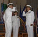 Three U.S. Navy officers in white dress uniforms are participating in a formal military ceremony, likely a change of command. Two officers are exchanging salutes while a third officer, a woman with captain or admiral insignia, stands between them as a presiding official. The ceremony takes place in a formal hall decorated with numerous military and state flags.